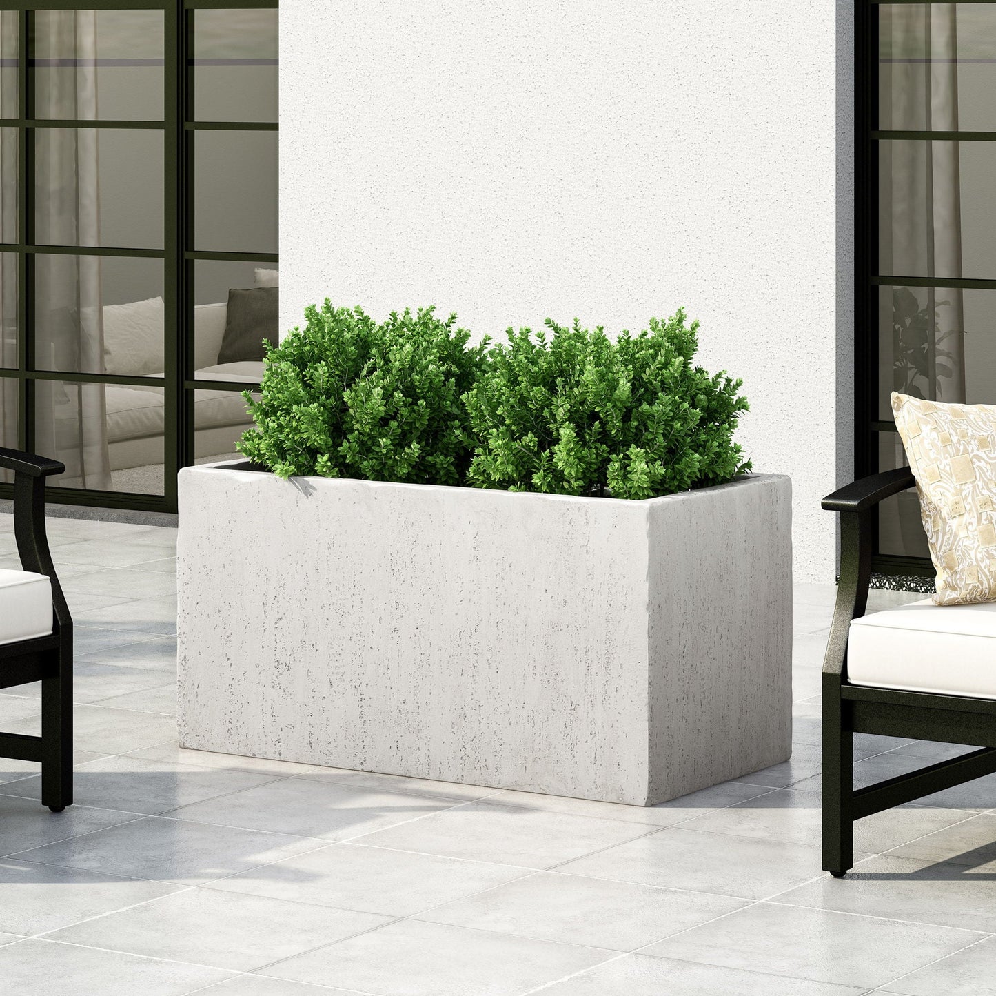 Rectangular planter with green plants on a patio with chairs and a white wall.