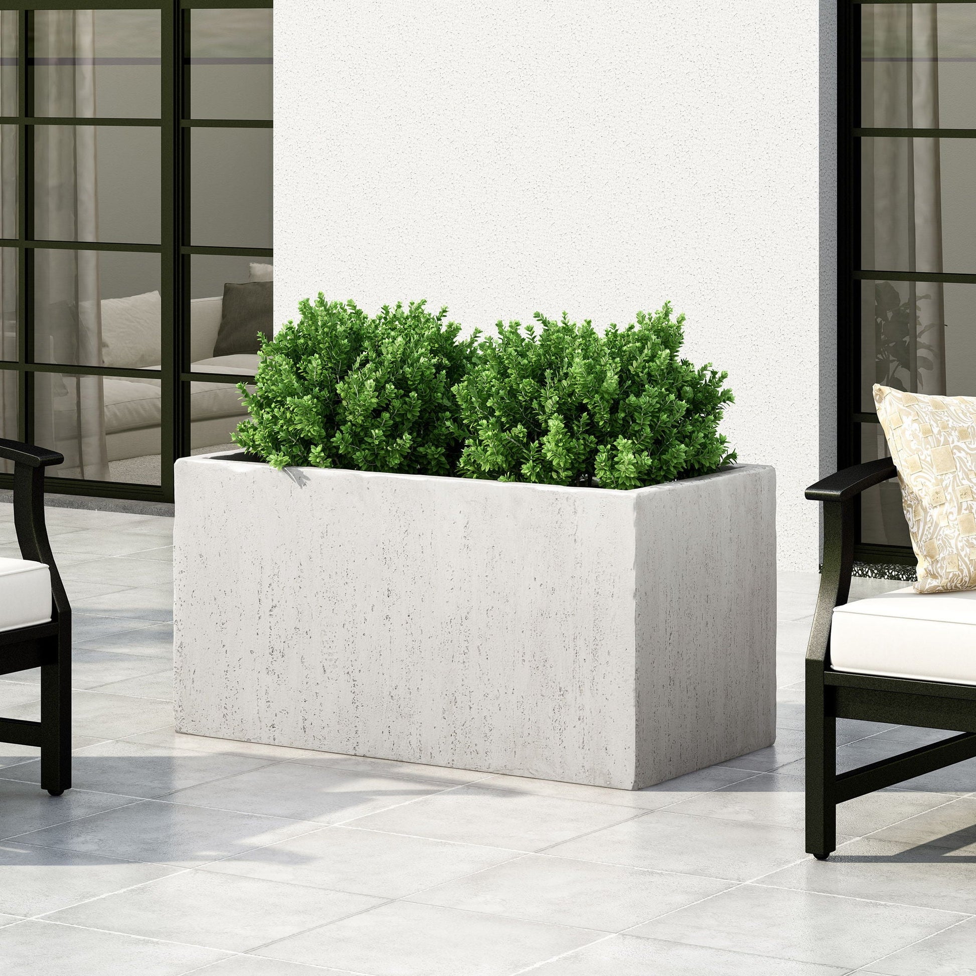 Rectangular planter with green plants on a patio with chairs and a white wall.