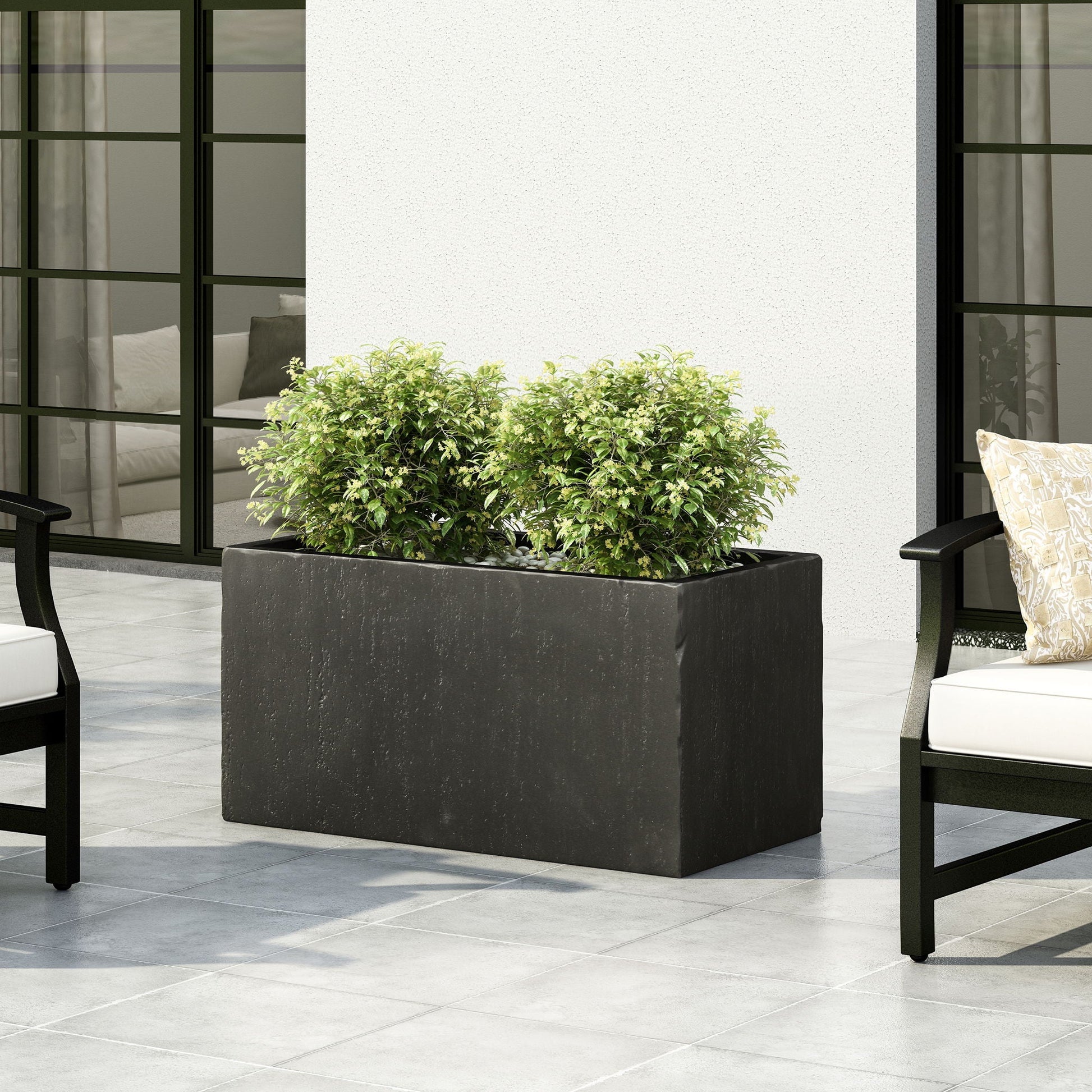 Black rectangular planter with green plants on a patio with chairs and cushions.