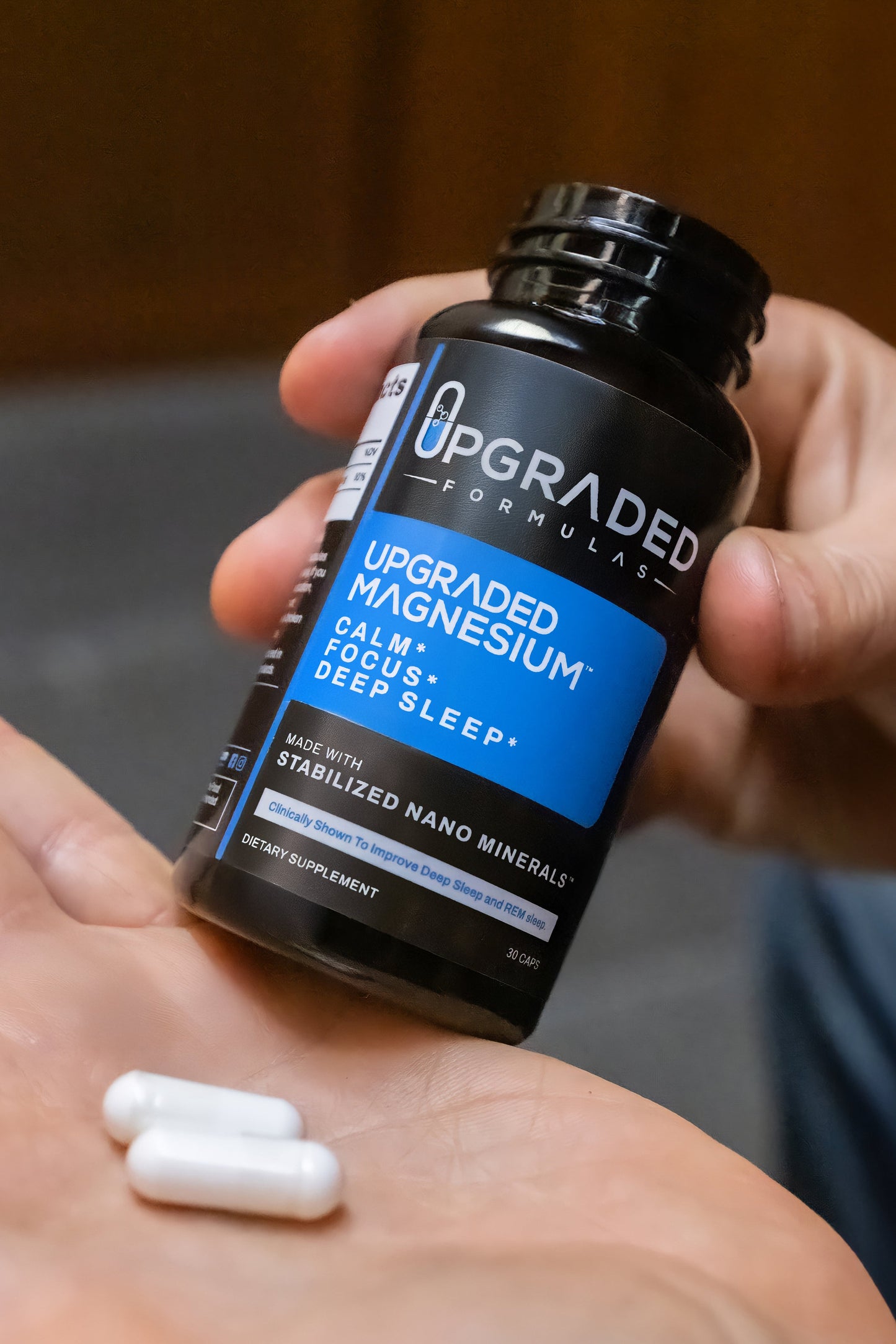 Hand holding a bottle of 'Upgraded Formulas' magnesium supplement with two capsules on an open palm.