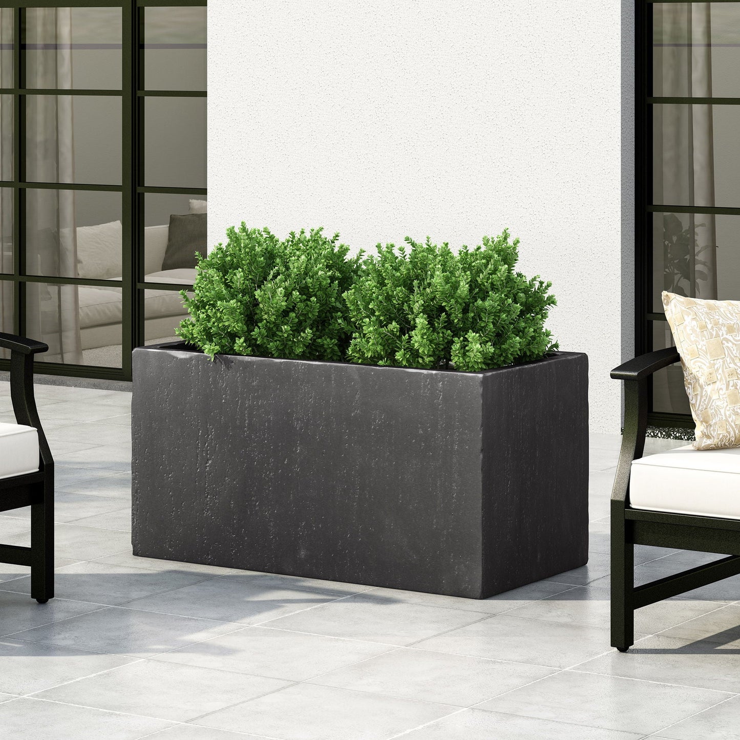 Black rectangular planter with green plants on a patio with chairs and a white wall.