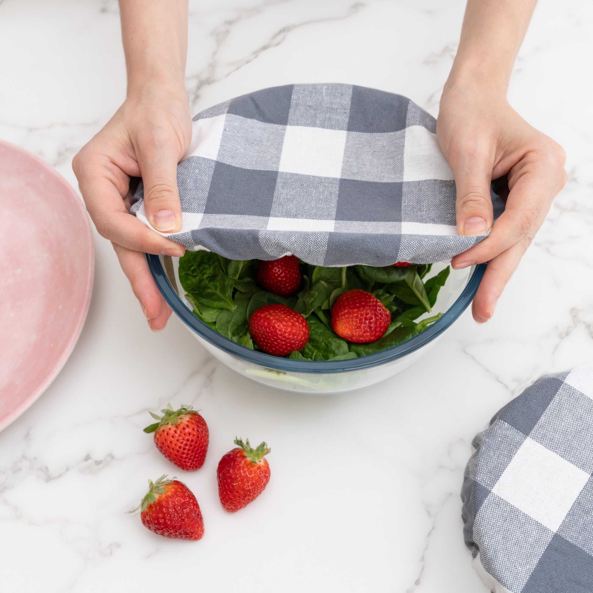 Person holding a bowl with a checkered cover, surrounded by strawberries on a marble surface.