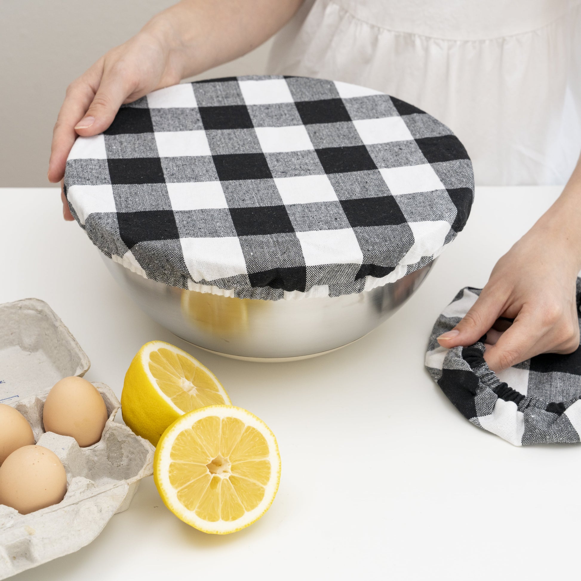 Person using a black and white checkered cloth to cover a metal bowl on a kitchen counter with lemons and eggs.
