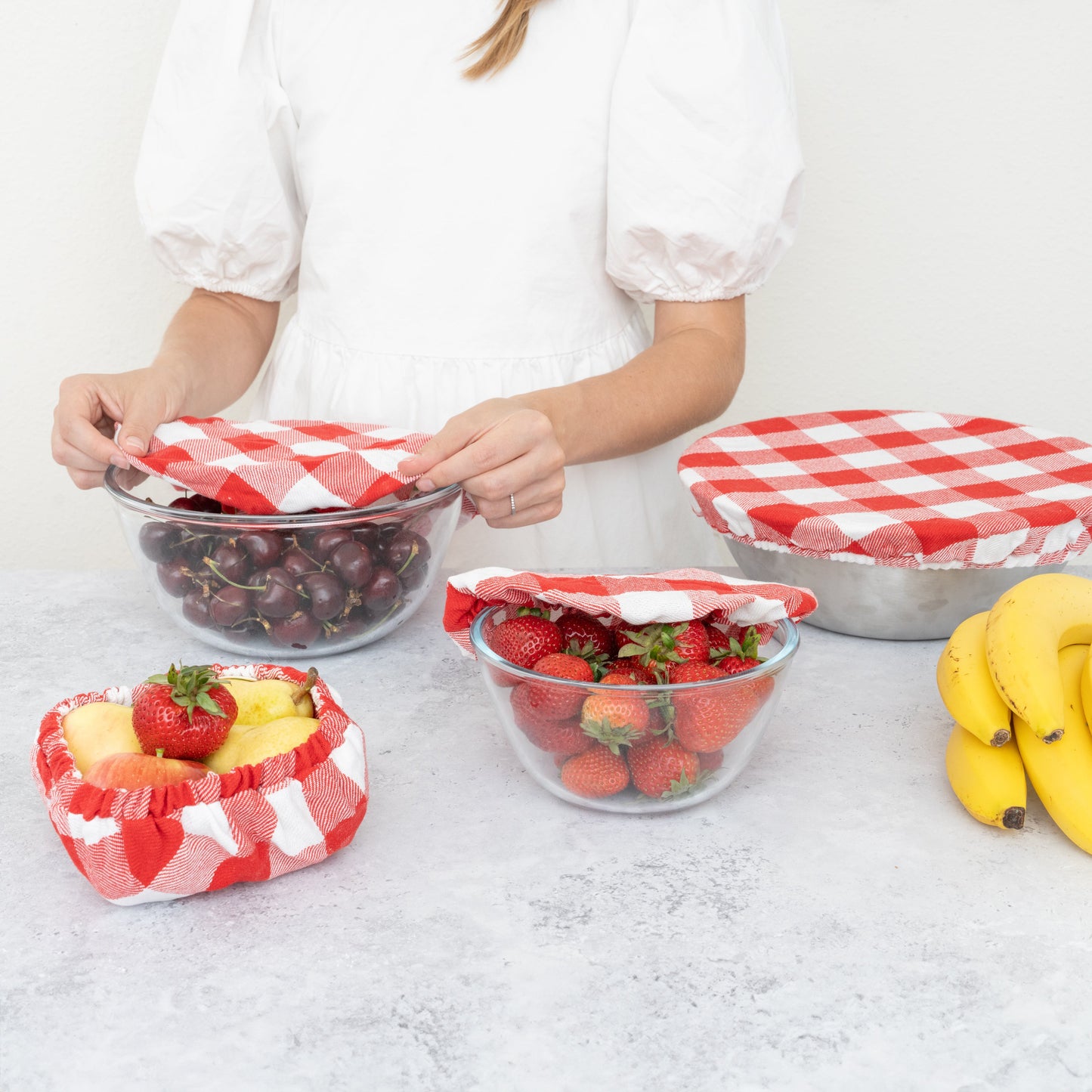 Person arranging fruits in glass bowls with red and white checkered covers on a light surface.