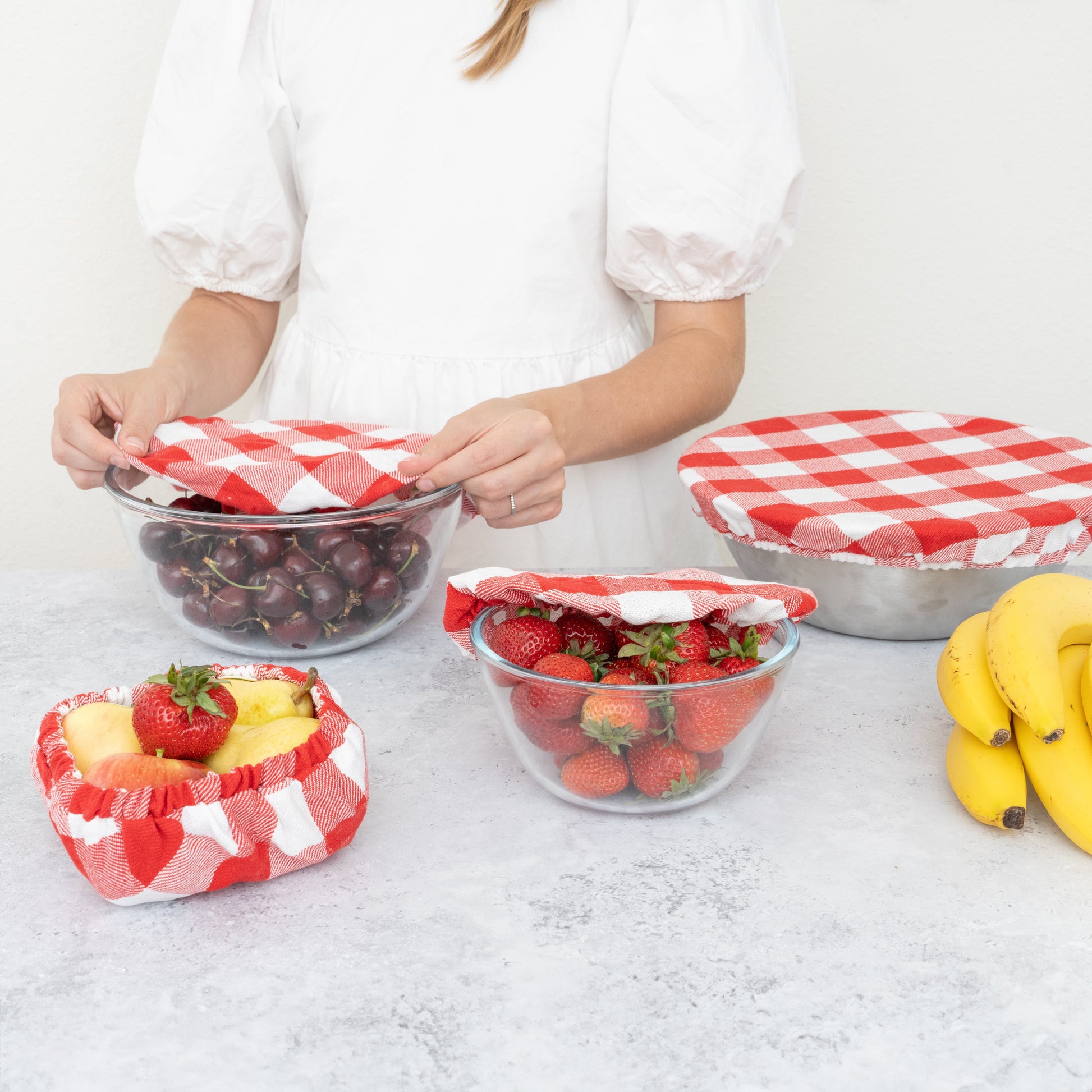 Person arranging fruits in glass bowls with red and white checkered covers on a light surface.