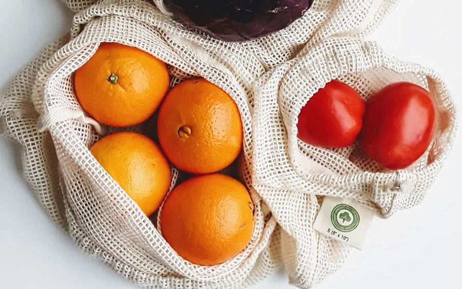 Fruits including oranges and tomatoes in reusable mesh bags on a white background